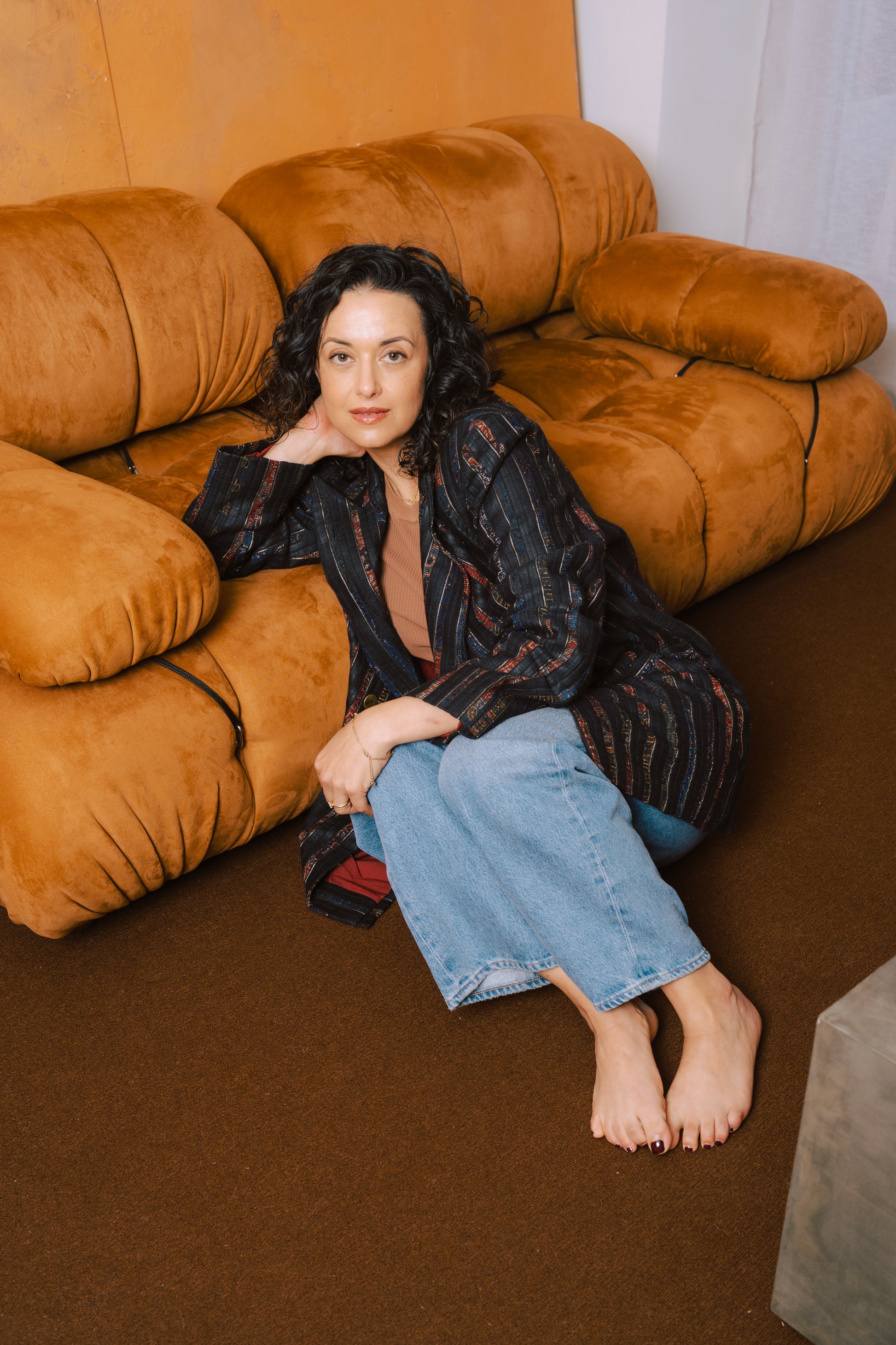 Woman sitting on a brown couch wearing a leather blazer jacket and blue jeans.