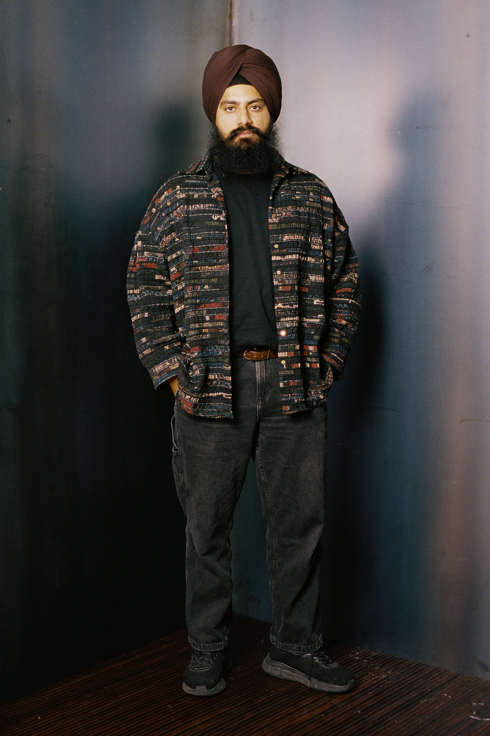 Man wearing a patterned shirt and turban against a plain background with black jeans and sneakers