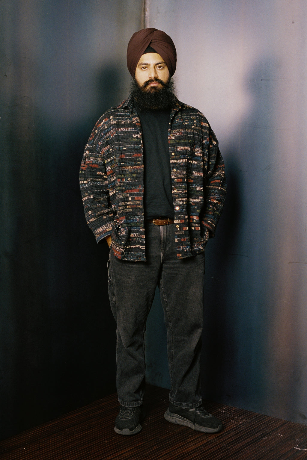 Man wearing a patterned shirt and turban against a plain background with black jeans and sneakers