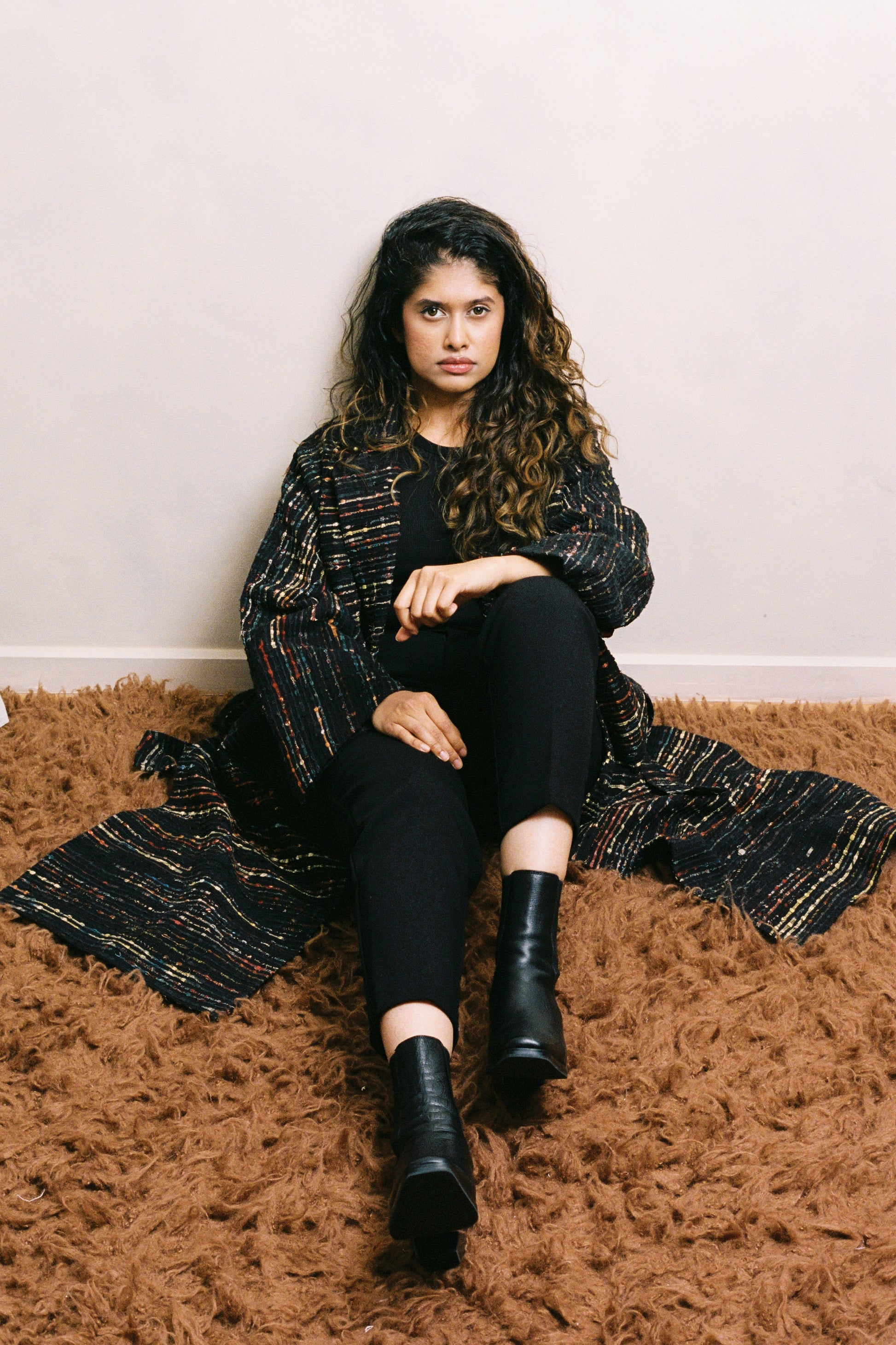 Woman sitting on a brown rug wearing a black and gold patterned coat, black pants, and black boots.