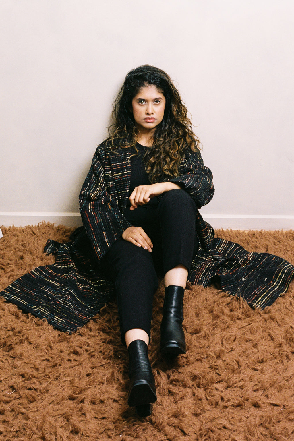 Woman sitting on a brown rug wearing a black and gold patterned coat, black pants, and black boots.