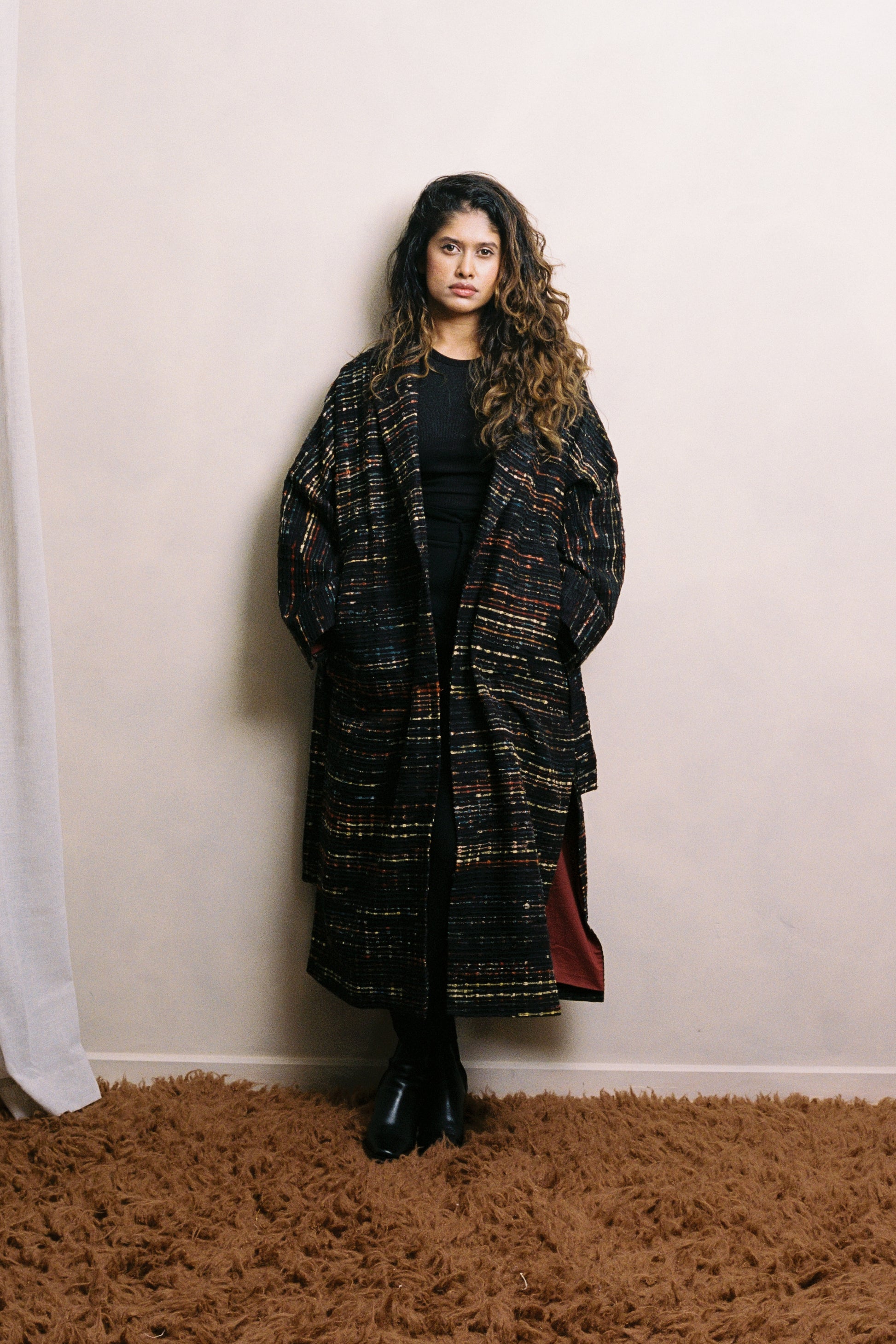 Woman wearing a long, patterned coat standing against a plain wall. She is in a relaxed pose. The coat features dropped shoulders, a soft, fluid silhouette, and a subtle taper through the body. Deep charcoal in tone, with ajrak detailing stitched. The styling conveys quiet elegance and ease.