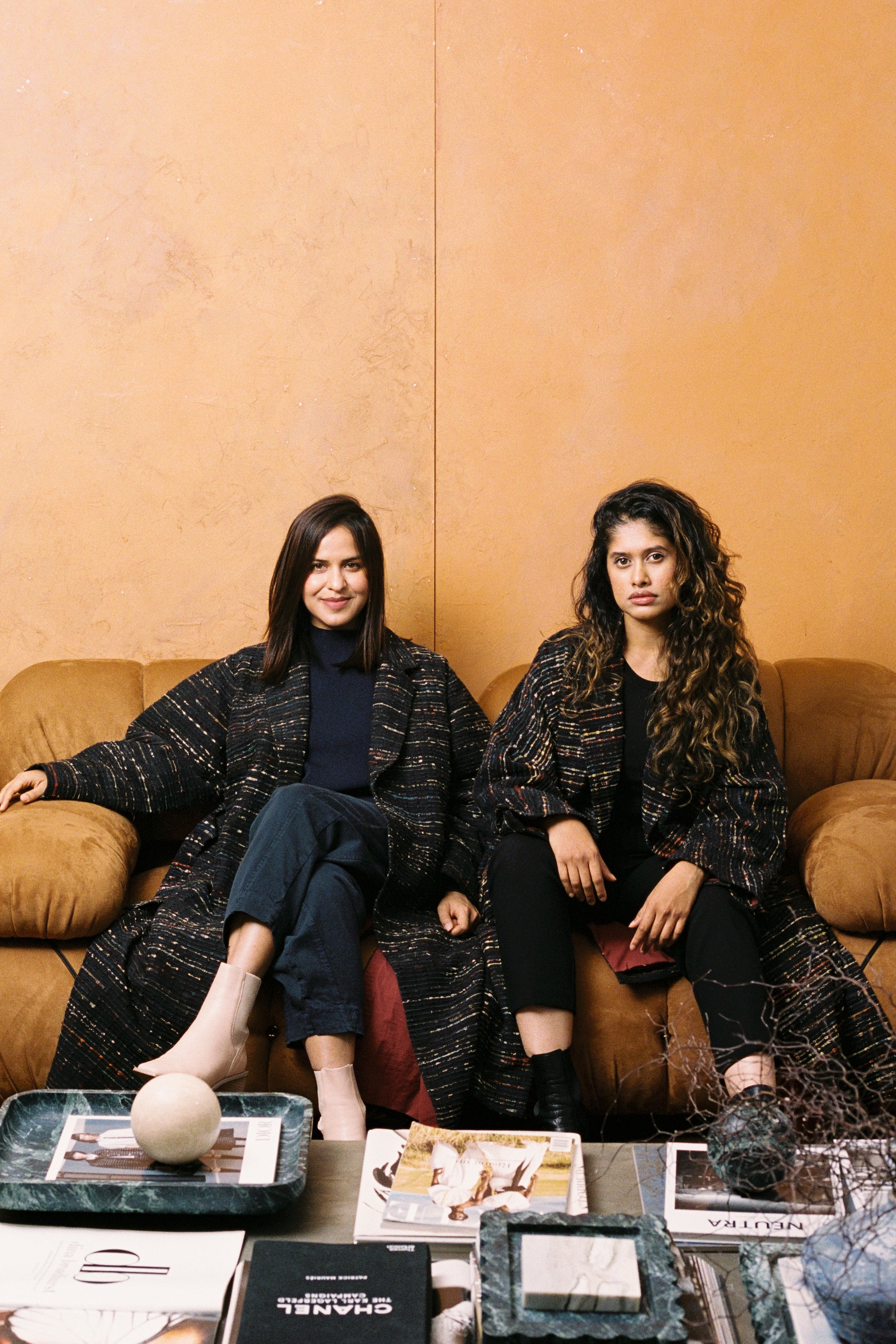 Two women sitting on a couch with a neutral background with black and brown boots
