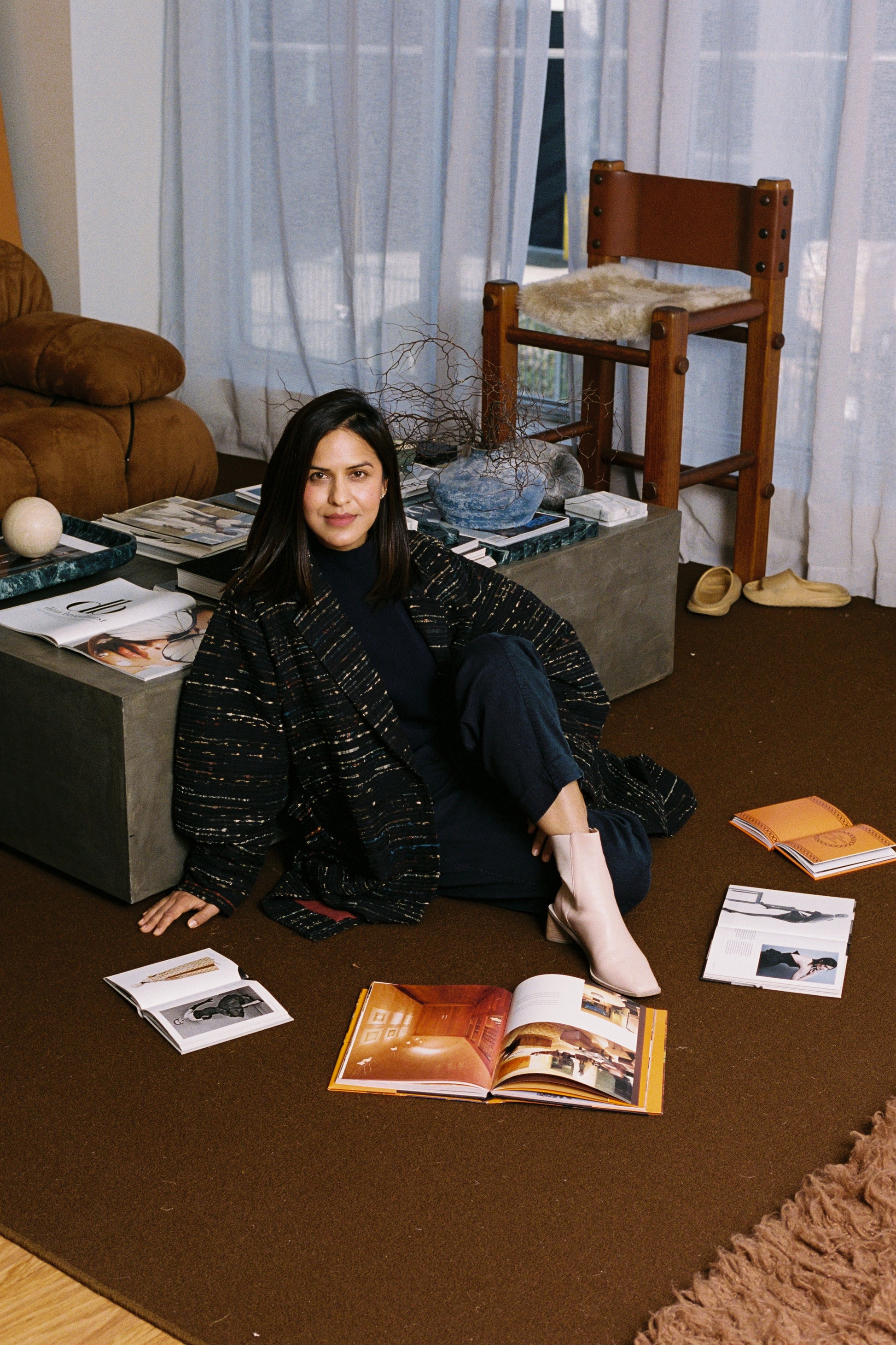 Woman sitting on the floor surrounded by books and magazines in a room with curtains and furniture.