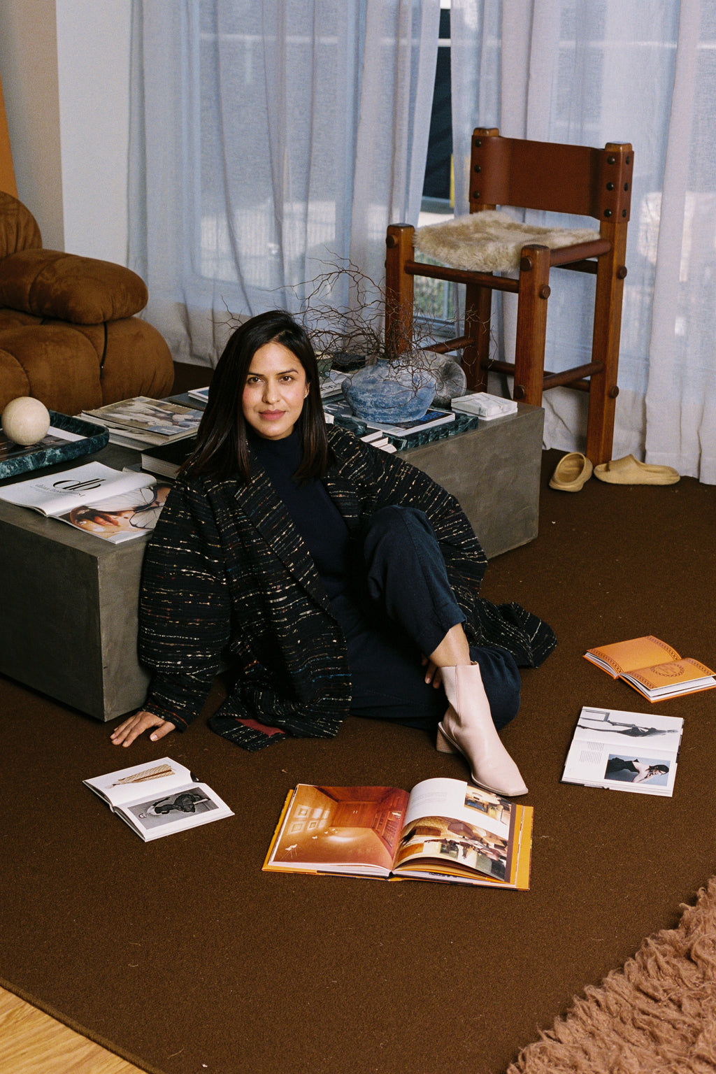 Woman sitting on the floor surrounded by books and magazines in a room with curtains and furniture.