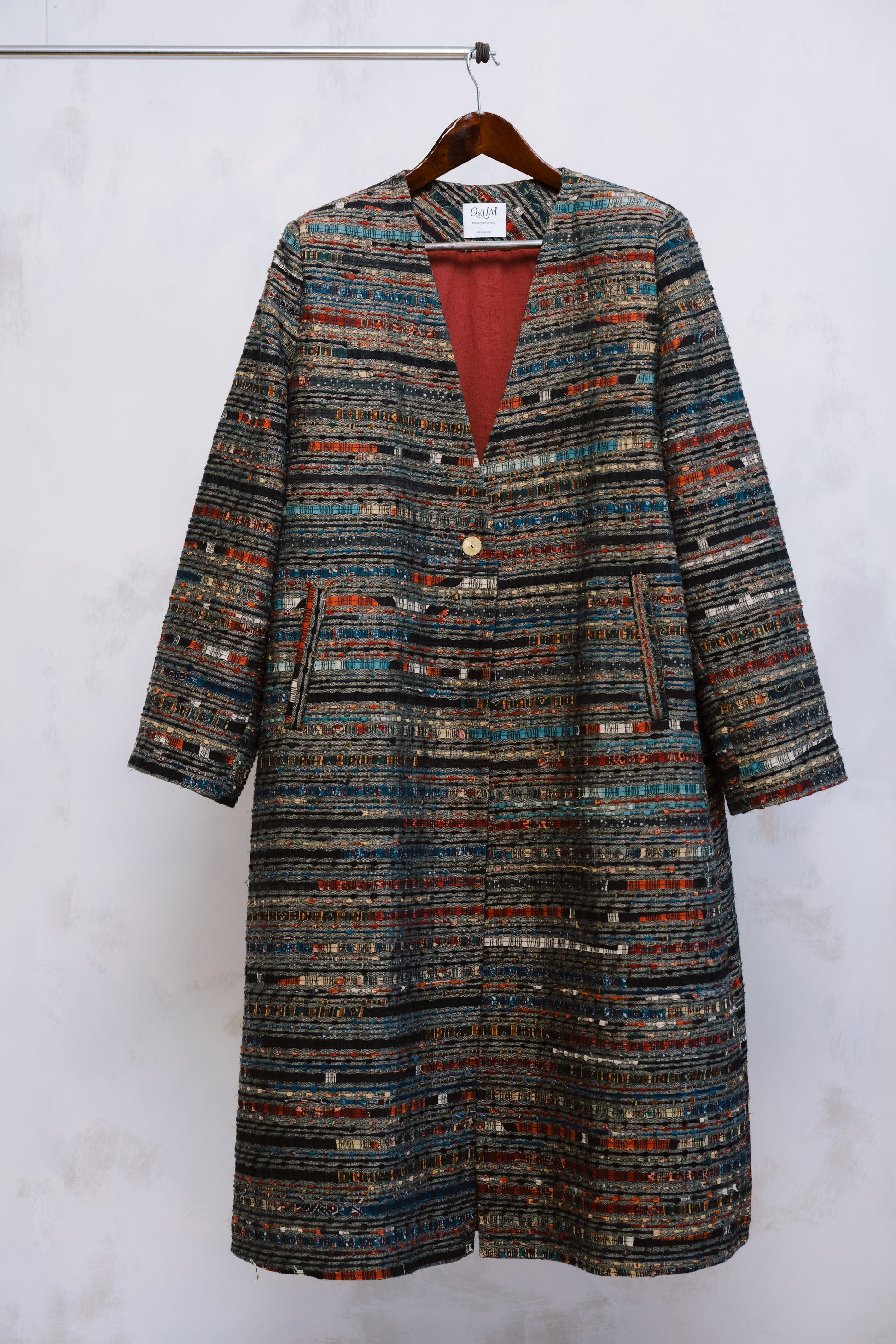 The Ash Coat on hanger, its minimalist silhouette contrasted by deep red ajrak lining - where structure meets story.