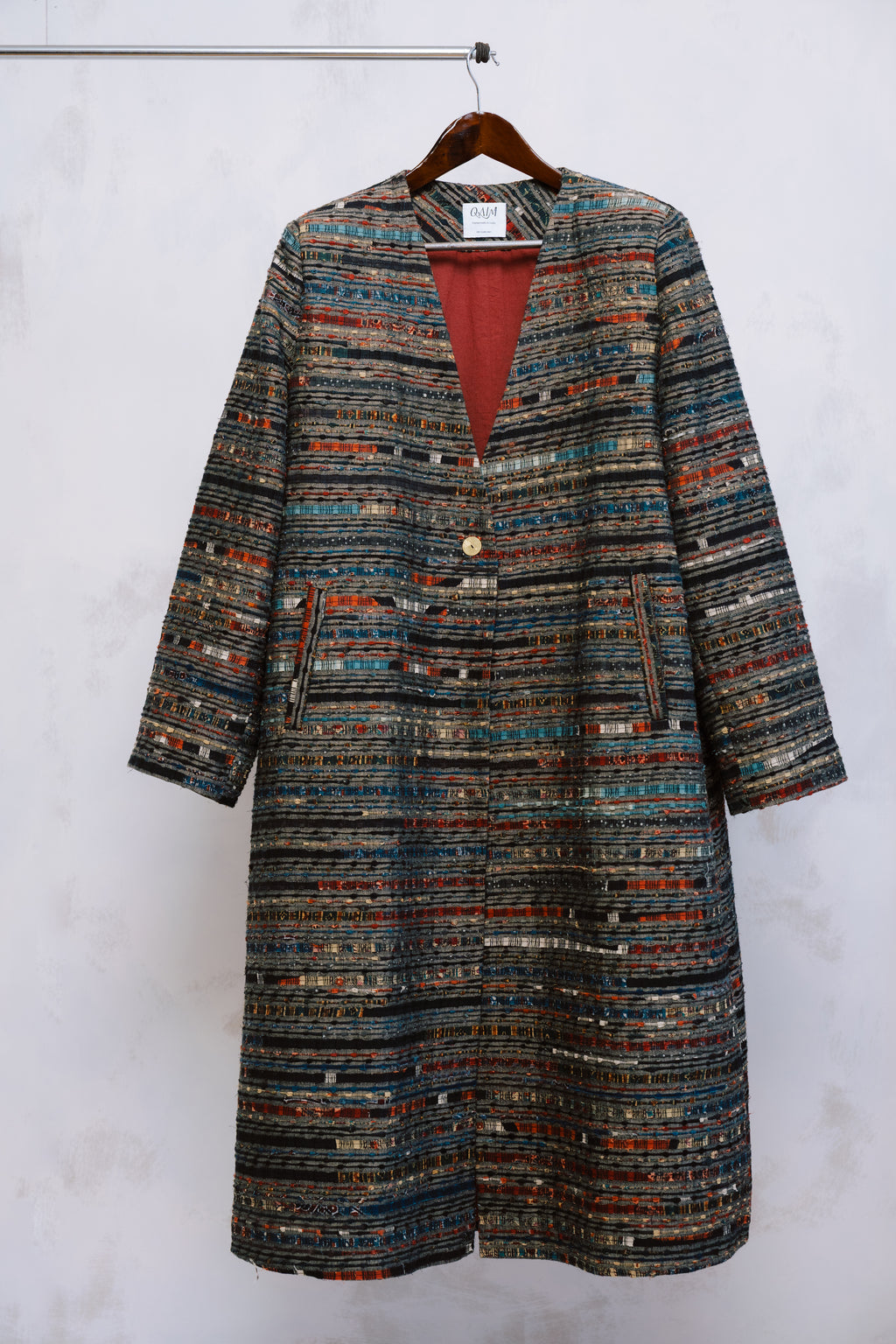 The Ash Coat on hanger, its minimalist silhouette contrasted by deep red ajrak lining - where structure meets story.